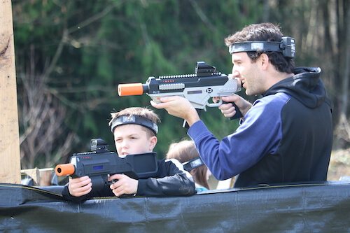 A smiling father and son take cover behind a barrier while playing laser tag.